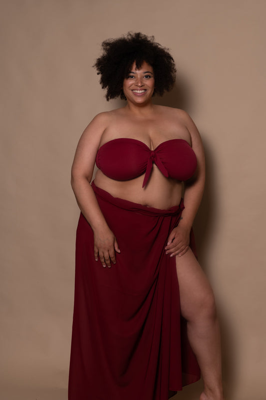 plussize woman wearing a pomegranate red bikini with pareo cover up against a beige background