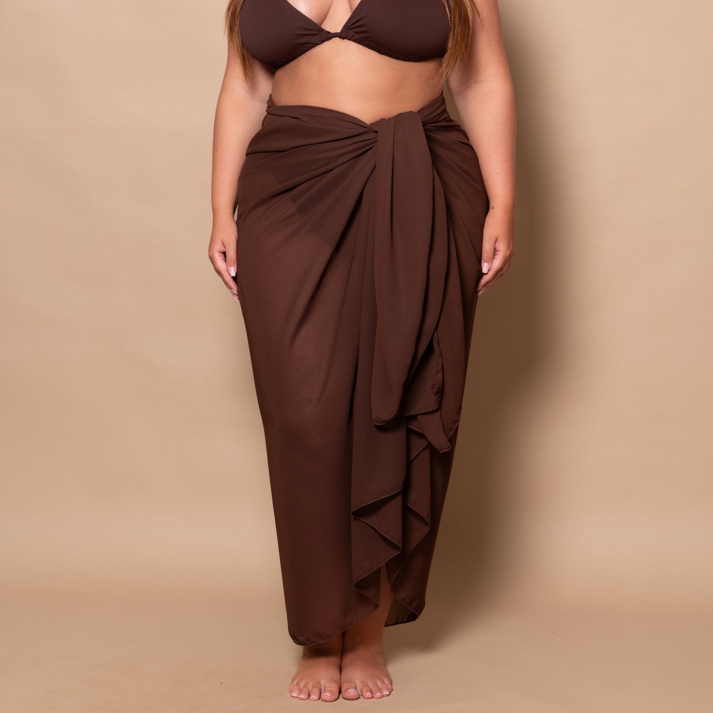 curvy woman wearing a chocolate brown bikini set and pareo cover up against a beige background