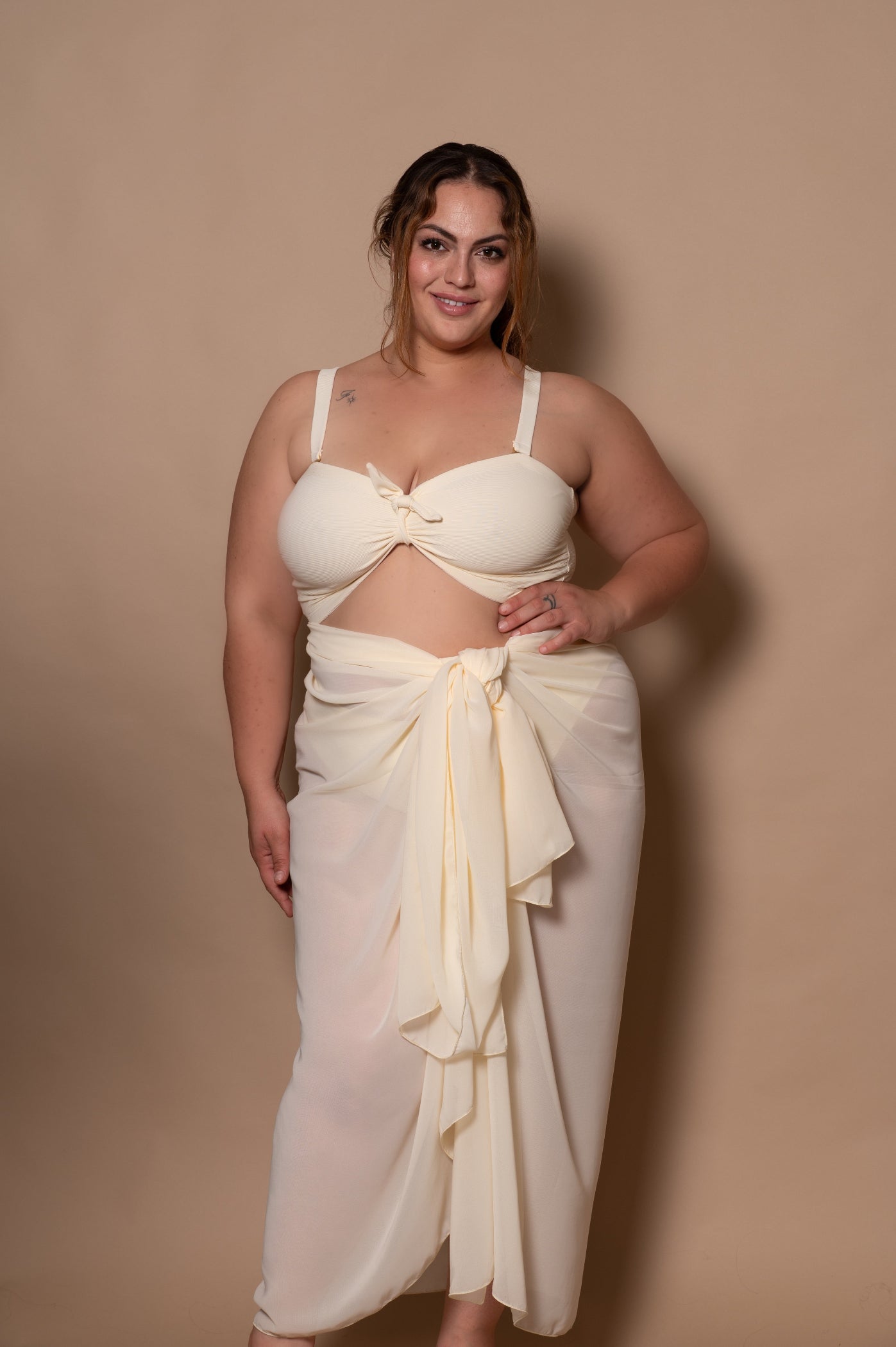 plussize woman wearing a white cream bikini with pareo cover up against a beige background