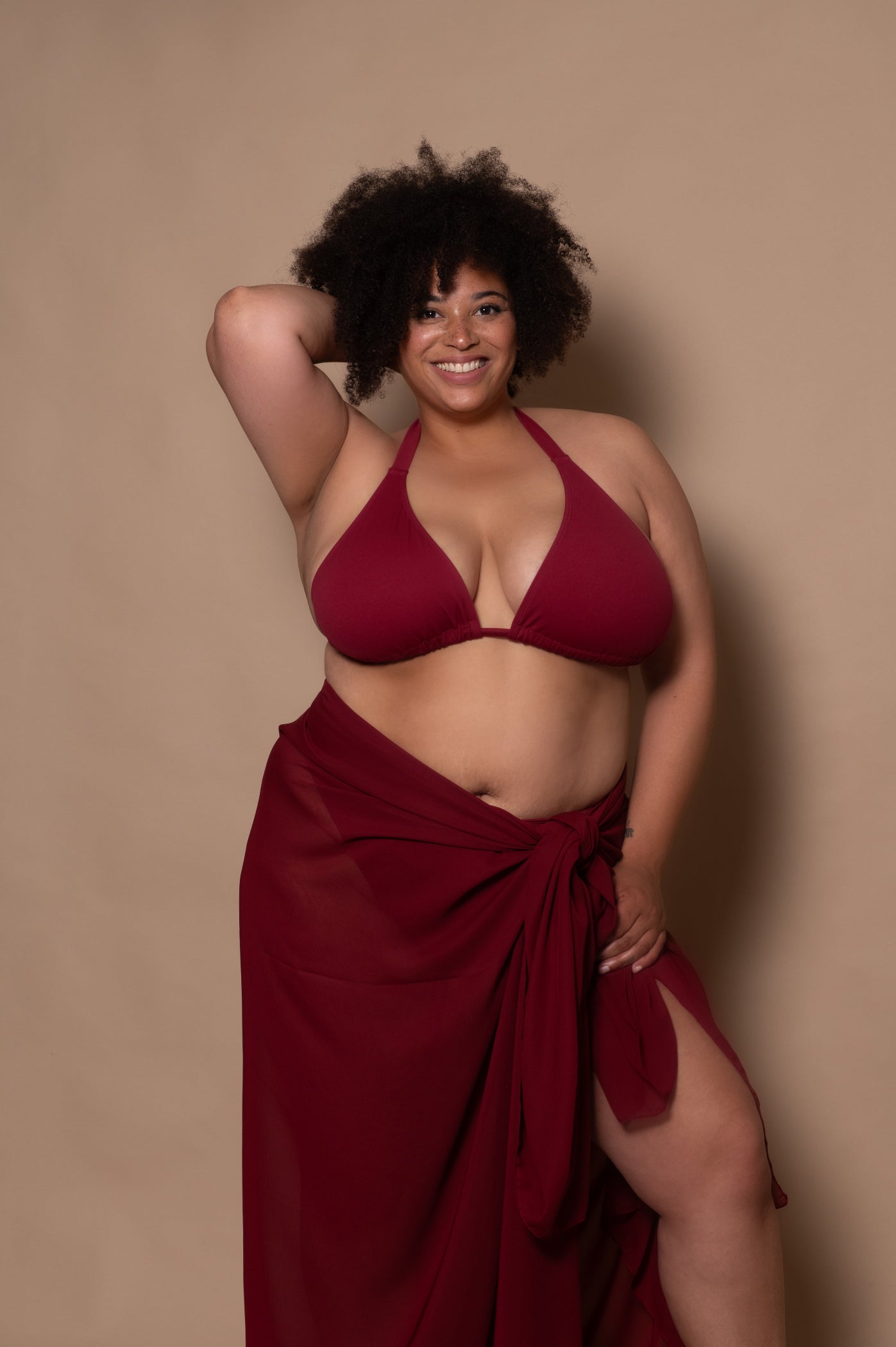 plussize woman wearing a pomegranate red bikini with pareo cover up against a beige background