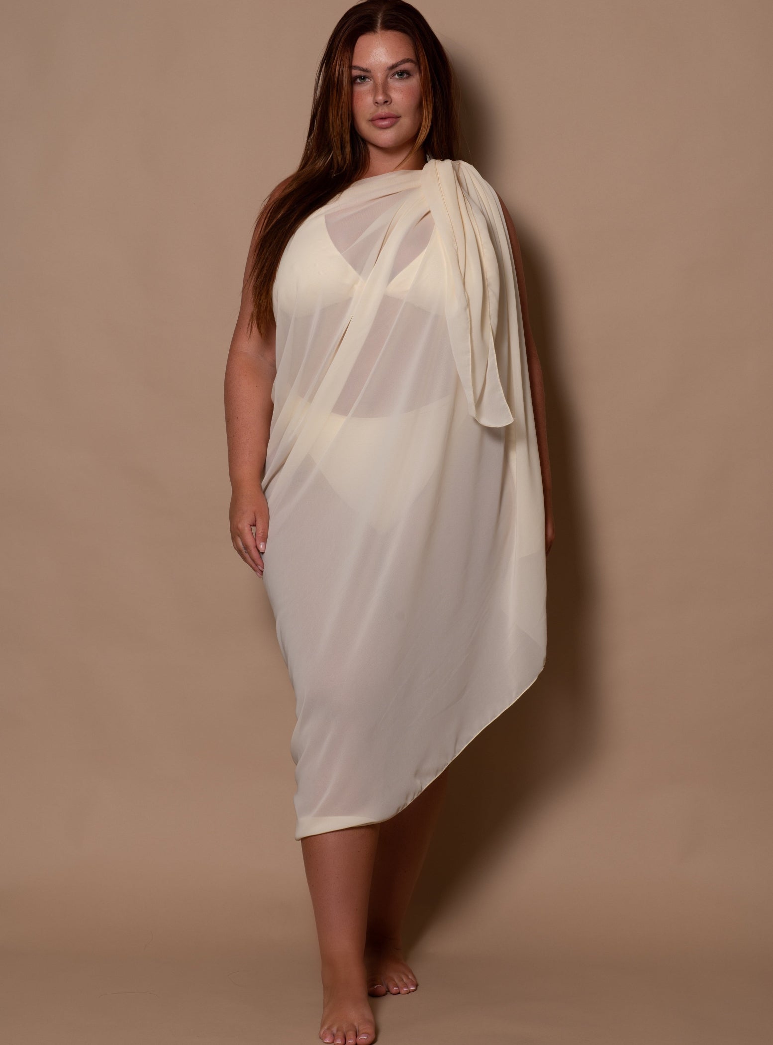 plussize woman wearing a white cream bikini top and white cream sarong against a brown background