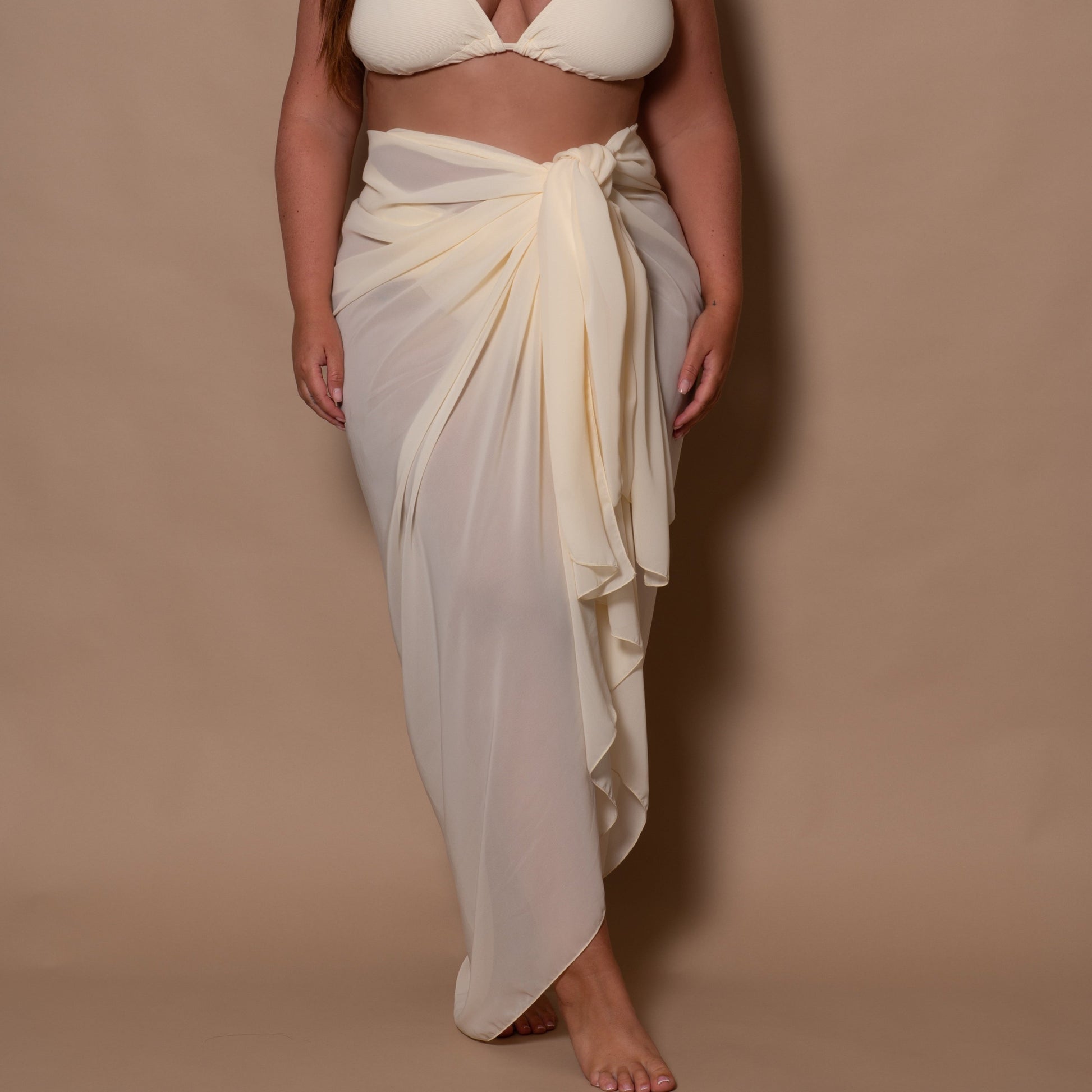 plussize woman wearing a white cream bikini top and white cream sarong against a brown background