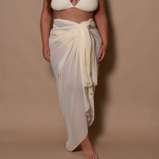 plussize woman wearing a white cream bikini top and white cream sarong against a brown background