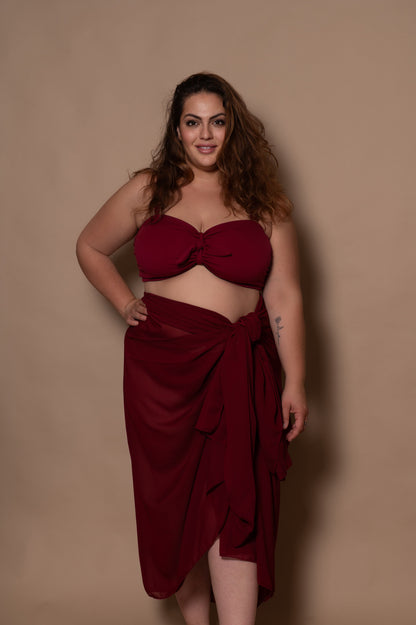 plussize woman wearing a pomegranate red bikini with pareo cover up against a beige background